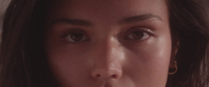 Nahaufnahme des Gesichts einer Frau mit langen dunklen Haaren, die direkt in die Kamera schaut. Ihre Haut ist glatt, sie ist minimal geschminkt und trägt einen kleinen goldenen Ohrring im linken Ohr. Der Hintergrund ist neutral.