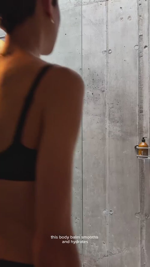 Eine Person in einem schwarzen BH steht vor einer Badezimmerwand aus Beton mit einer Flasche Körperbalsam auf einem Regal. Der Text lautet: Dieser Körperbalsam glättet und spendet Feuchtigkeit.