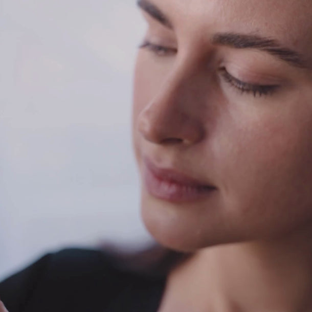 Nahaufnahme einer Frau mit geschlossenen Augen und ruhigem Gesichtsausdruck, die mit ihrer rechten Hand etwas außerhalb des Bildes hält. Der Hintergrund ist sanft verschwommen.