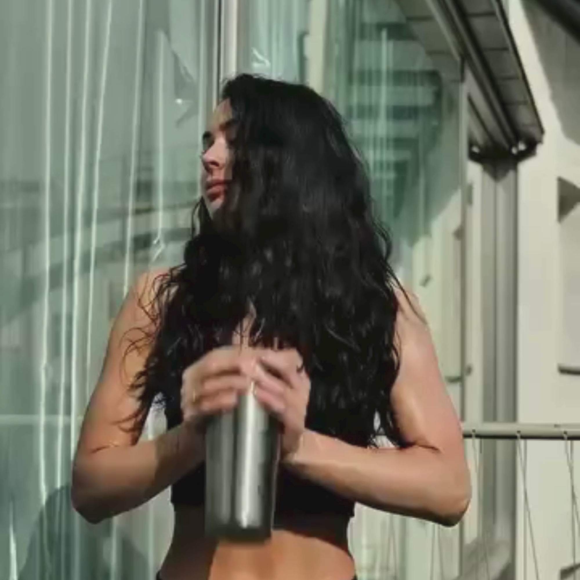 Eine Frau mit langen dunklen Haaren steht im Sonnenlicht auf einem Balkon, trägt einen schwarzen Sport-BH und hält mit beiden Händen eine metallene Shakerflasche. Sie blickt zur Seite, und hinter ihr sind Glasscheiben zu sehen.