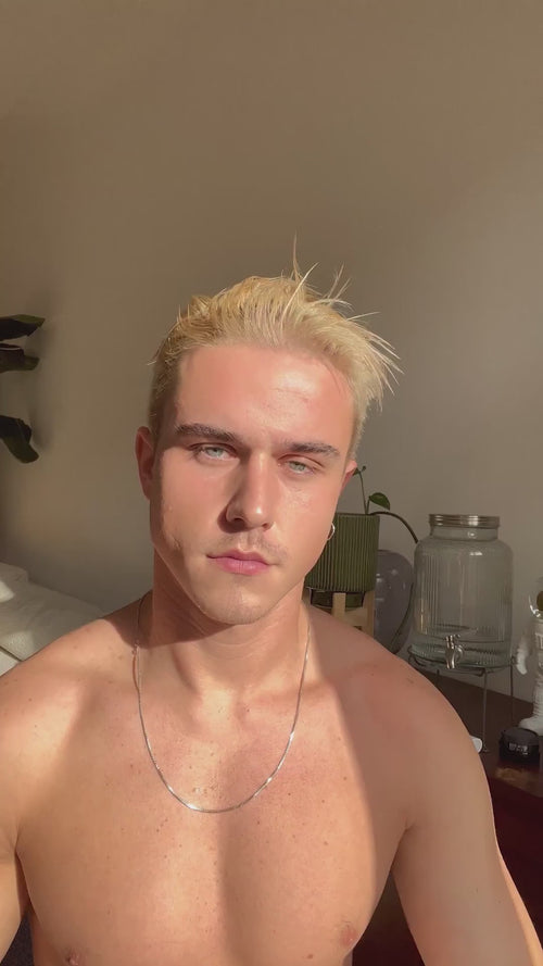 Eine Person, die einen schwarzen Ärmel trägt, hält eine kleine Flasche mit einer Flüssigkeit hoch und versprüht sie, während hinter ihr ein durchsichtiger Vorhang und ein verschwommener Hintergrund zu sehen sind.