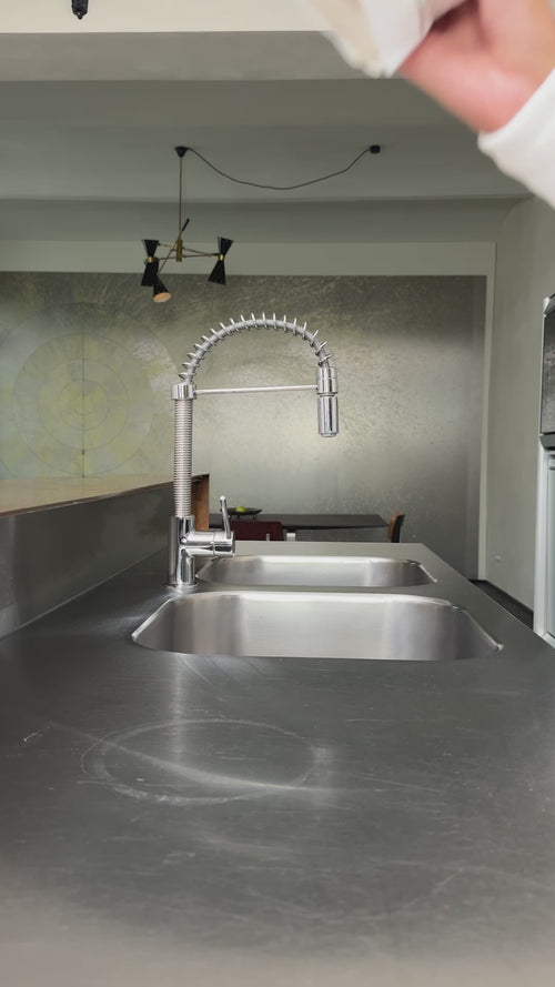 Eine moderne Küche mit einer Arbeitsplatte aus rostfreiem Stahl, einer Doppelspüle und einem hohen, flexiblen Wasserhahn. Im Hintergrund sind eine dekorative Wand und Pendelleuchten zu sehen. Oben rechts ist die bandagierte Hand einer Person teilweise sichtbar.
