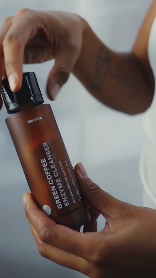 Jemand öffnet eine braune Flasche mit der Aufschrift Daluma GREEN COFFEE CLEANSER ENZYMPEELING und hält sie in der einen Hand, während er mit der anderen den schwarzen Verschluss abschraubt.