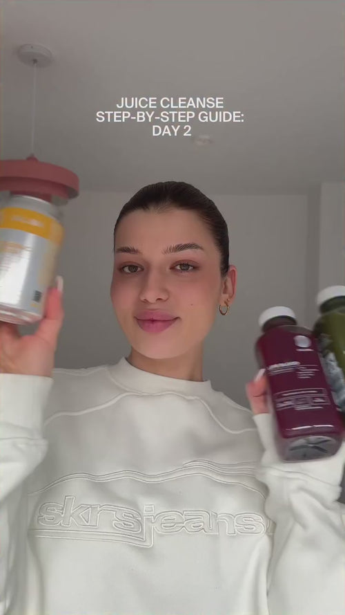 Eine junge Frau in einem weißen Sweatshirt hält drei Daluma ALKALISCHE SAFTKUR - 3 TAGE Flaschen. Über ihr: JUICE CLEANSE SCHRITT-FÜR-SCHRITT-ANLEITUNG: DAY 2. Sie lächelt sanft in einem hellen, minimalistischen Raum.