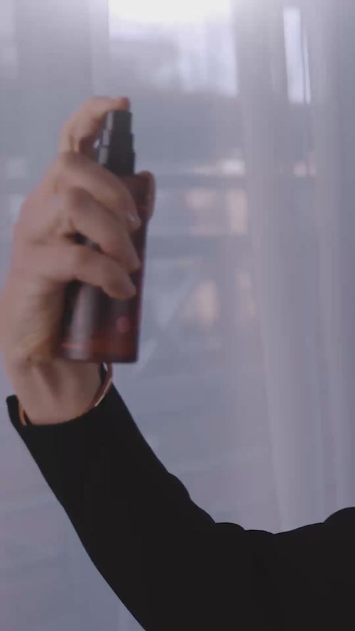 Eine Person, die einen schwarzen Ärmel trägt, hält eine kleine Flasche mit einer Flüssigkeit hoch und versprüht sie, während hinter ihr ein durchsichtiger Vorhang und ein verschwommener Hintergrund zu sehen sind.