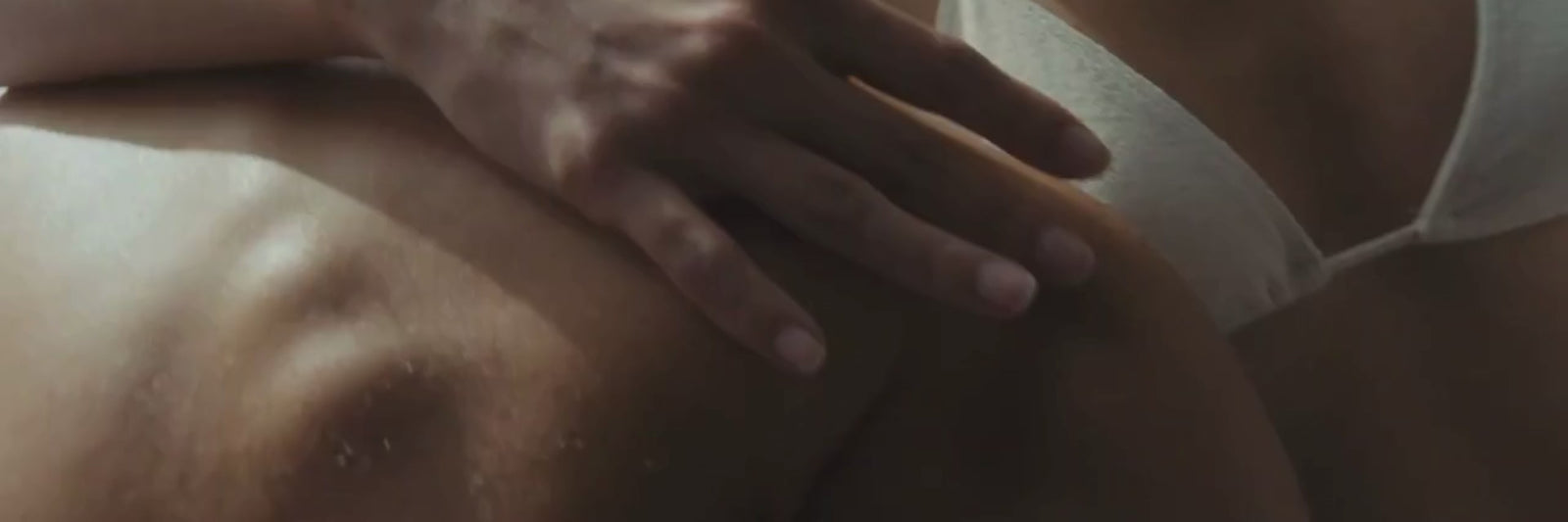 Eine Nahaufnahme einer Hand, die sanft auf dem nackten Rücken einer Person ruht, neben der eine andere Person mit einem weißen BH zu sehen ist. Das Bild vermittelt eine intime, weiche Atmosphäre.