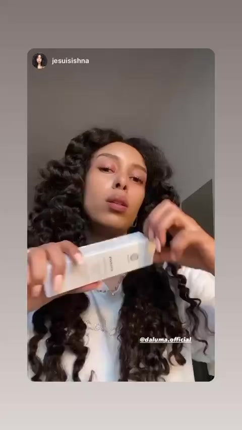 Eine Frau mit langem, lockigem Haar hält eine weiße Kosmetikflasche in der Hand und untersucht sie. Sie trägt ein weißes Oberteil und blickt leicht nach oben. Das Instagram-Handle @daluma.official ist auf dem Bild zu sehen.