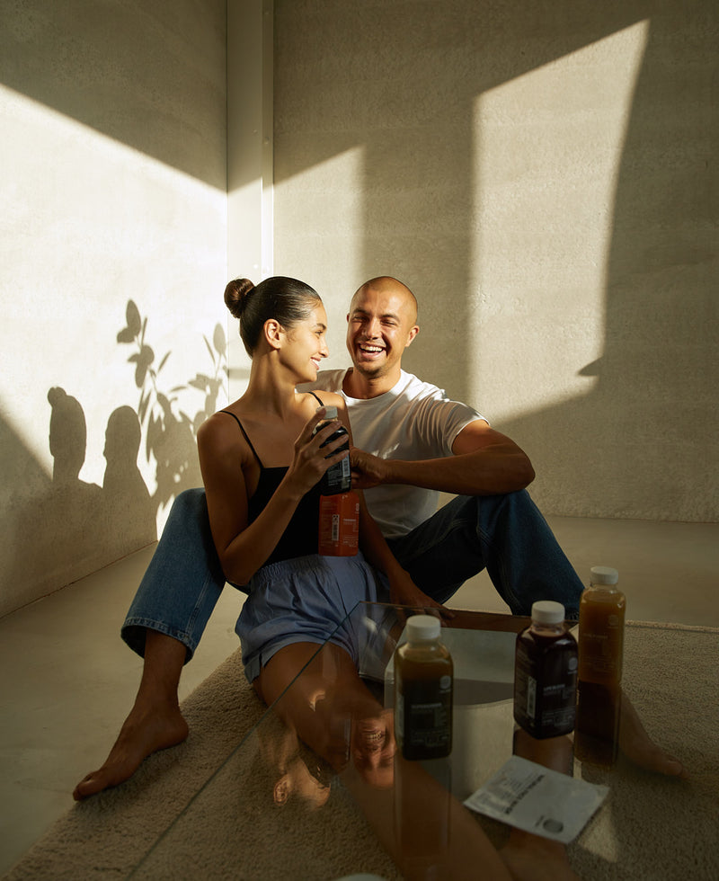 Ein lächelndes Paar sitzt auf dem Boden an einer sonnenbeschienenen Wand und teilt sich Getränke in Flaschen. Der Mann trägt ein weißes Hemd und Jeans, die Frau ein schwarzes Tanktop und Shorts. Weitere Flaschen und Päckchen stehen auf einem Glastisch vor ihnen.