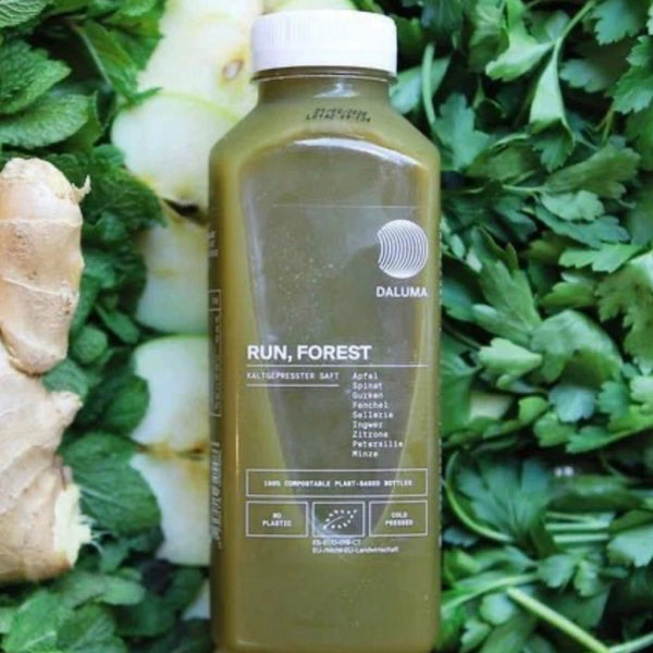 Eine grüne Saftflasche mit der Aufschrift DALUMA RUN, FOREST steht auf einem Bett aus frischem Grün, darunter Petersilie und Ingwerwurzel. Die transparente Flasche zeigt die grüne Flüssigkeit im Inneren und listet die pflanzlichen Inhaltsstoffe auf dem Etikett auf.