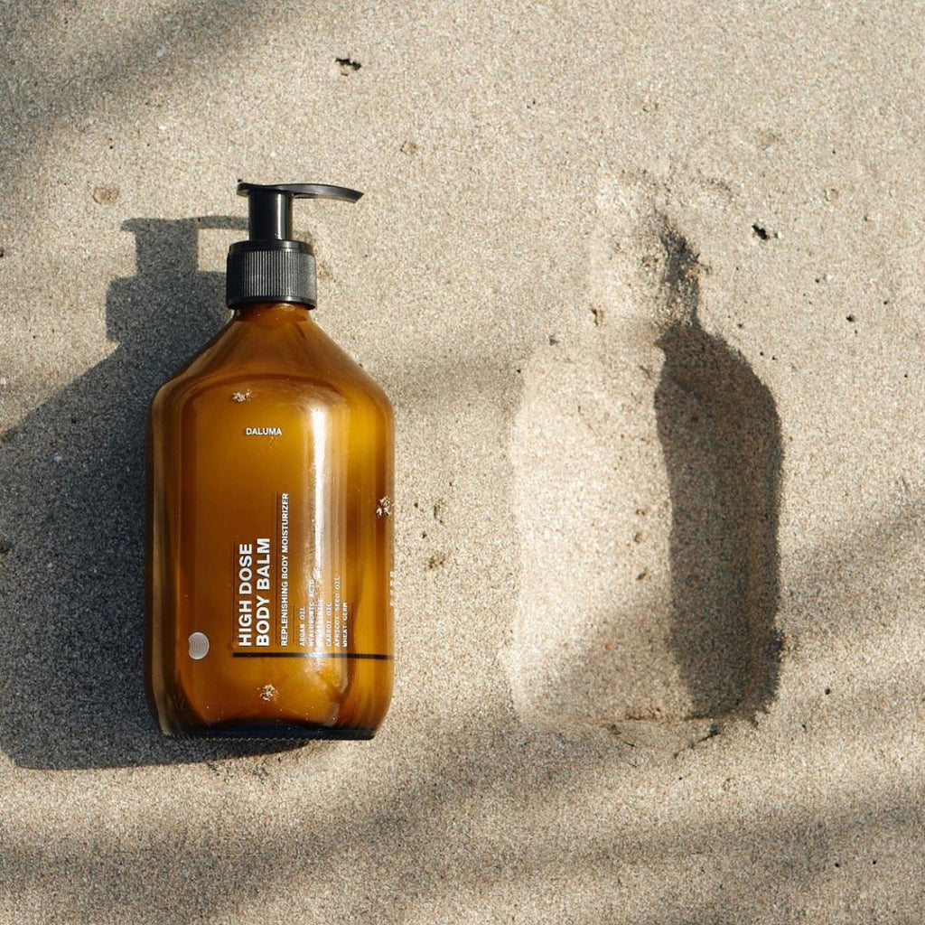 Eine braune Pumpflasche mit der Aufschrift SKIN NOURISHING DUO von DALUMA ruht neben ihrem Abdruck auf dem Sand und erinnert an ihre 6-Elemente-Hautreparaturformel, während sich sanfte Schatten über die Szene legen.