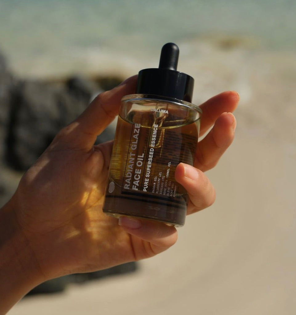 Eine Hand hält eine Flasche DALUMA HOLISTIC SUMMER GLOW Gesichtsöl mit einer Pipette und einer klaren, bernsteinfarbenen Flüssigkeit, ideal für die Hautreparatur, vor einem verschwommenen Strandhintergrund mit Sand und Felsen.