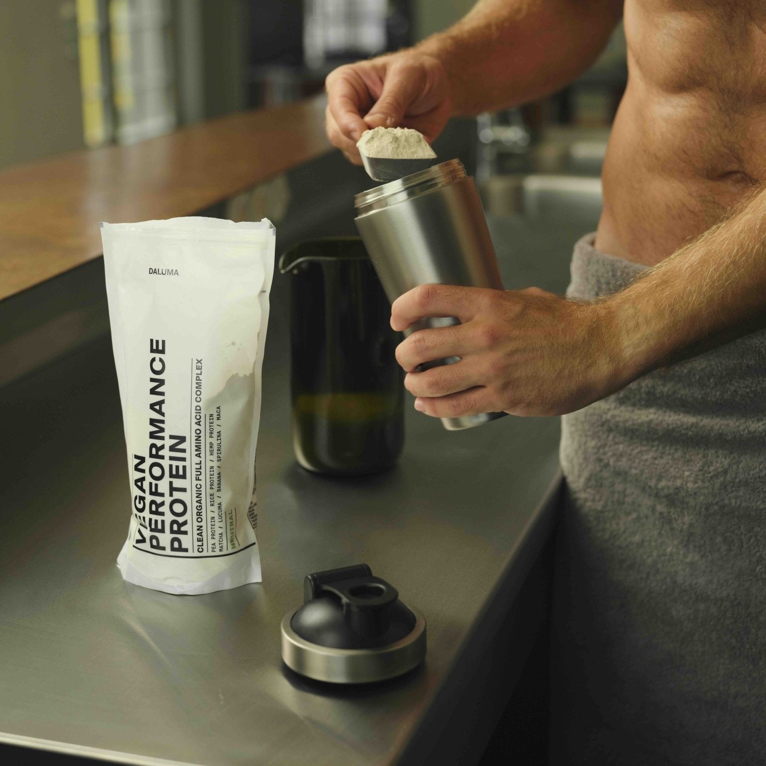 Nach dem Training gibt eine mit einem Handtuch bekleidete Person veganes Proteinpulver in den DALUMA PROTEIN SHAKER EDELSTAHL auf der Küchentheke und entscheidet sich für diese nachhaltige Alternative mit einem Performance Protein Beutel in der Nähe.