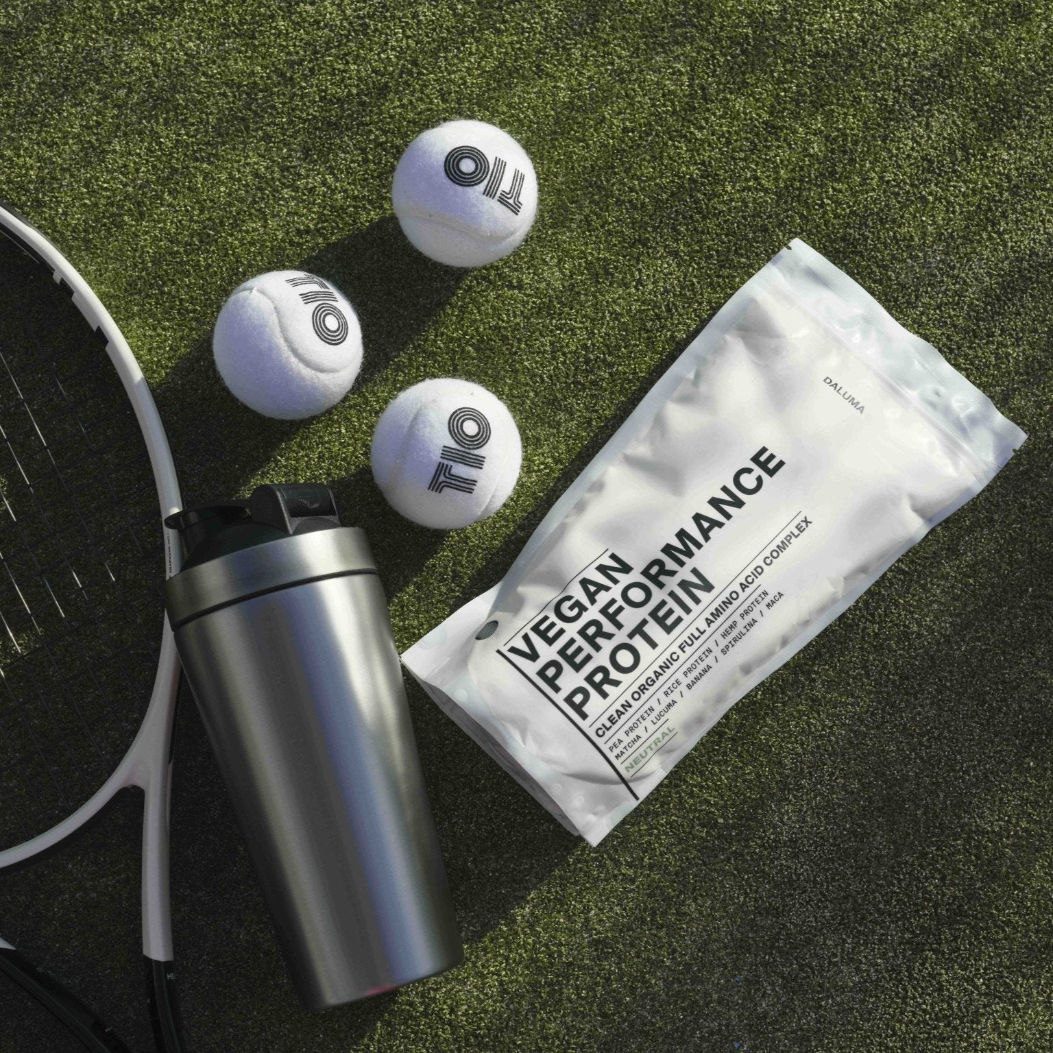 Ein DALUMA PROTEIN SHAKER EDELSTAHL, ein Vegan Performance Protein Beutel, drei Tennisbälle und ein Tennisschläger liegen auf grünem Kunstrasen - eine nachhaltige Wahl für sportliche Höchstleistungen.