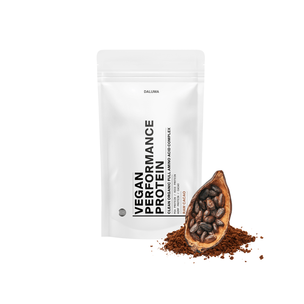 Ein weißer DALUMA VEGANES PERFORMANCE PROTEIN KAKAO-Beutel mit schwarzer Schrift und wiederverschließbarem Deckel steht aufrecht neben einer mit Bohnen und rohem Kakao gefüllten Kakaoschote und hebt den pflanzlichen Eiweißgehalt in einer minimalistischen Verpackung hervor.