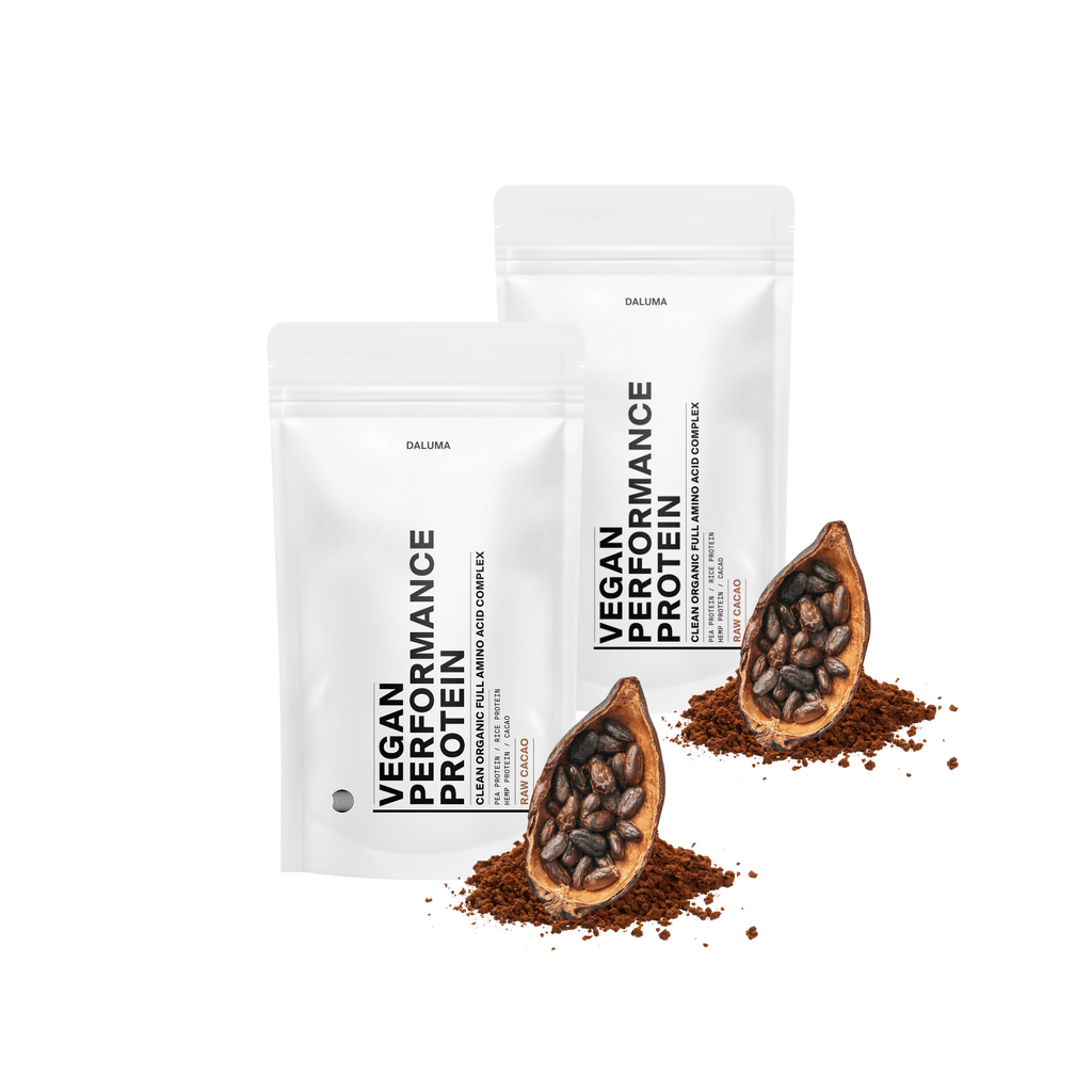 Zwei weiße DALUMA VEGANES PERFORMANCE PROTEIN KAKAO DOUBLE-Beutel stehen aufrecht, mit Kakaopulver, Rohkakao und Kakaoschoten vor jedem Beutel vor einem weißen Hintergrund.