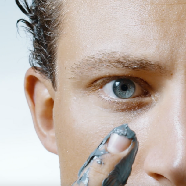 Nahaufnahme des Gesichts einer Person mit blauen Augen, nassem Haar und grauem Lehm oder Schlamm an den Fingern, den sie auf ihre Wange aufträgt. Der Hintergrund ist einfarbig und hell.